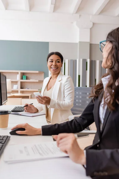 Happy multiracial businesswoman with smartphone looking at colleague working on blurred foreground — Stock Photo