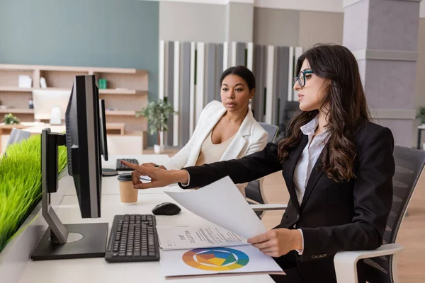 Manager in eyeglasses holding paper and pointing at computer monitor near multiracial colleague - foto de stock