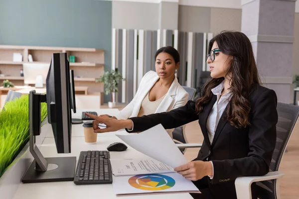 Manager in eyeglasses holding paper and pointing at computer monitor near multiracial colleague — Photo de stock