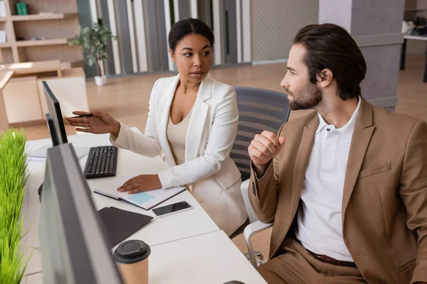 Multiracial businesswoman pointing at computer monitor and talking to colleague in office — Stock Photo