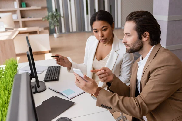 Thoughtful businessman pointing at smartphone while working with multiracial colleague in office — Stock Photo