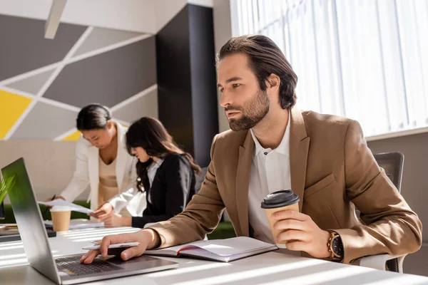 Focused businessman with coffee to go using laptop near interracial colleagues working on blurred background — Stock Photo