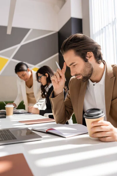 Thoughtful businessman with coffee to go looking in notebook near interracial colleagues working on blurred background — Stock Photo