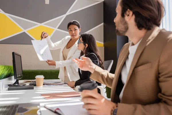 Multiracial woman showing blueprint to colleagues near desk with computer and documents on blurred foreground — Stock Photo
