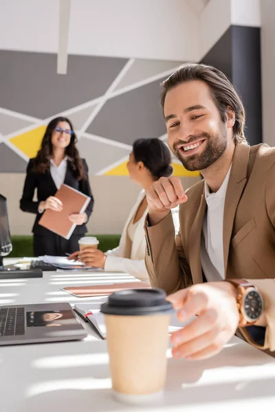 Smiling manager reaching takeaway drink while interracial businesswomen talking on blurred background — Stock Photo