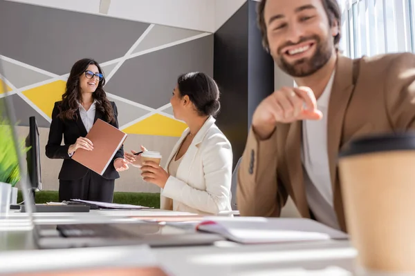 Interracial businesswomen with folder and coffee to go talking in office near manager smiling on blurred foreground — Stock Photo