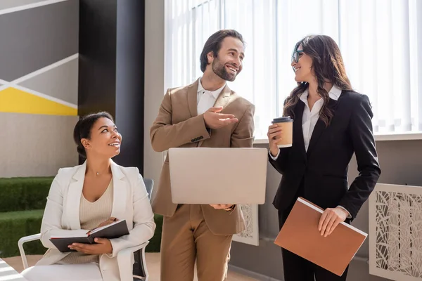 Smiling manager with laptop talking to colleague standing with coffee to go and folder near multiracial businesswoman in office — Stock Photo