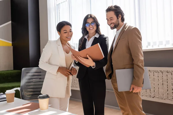 Happy businesswoman in eyeglasses holding folder with documents near smiling multiethnic colleagues in office - foto de stock