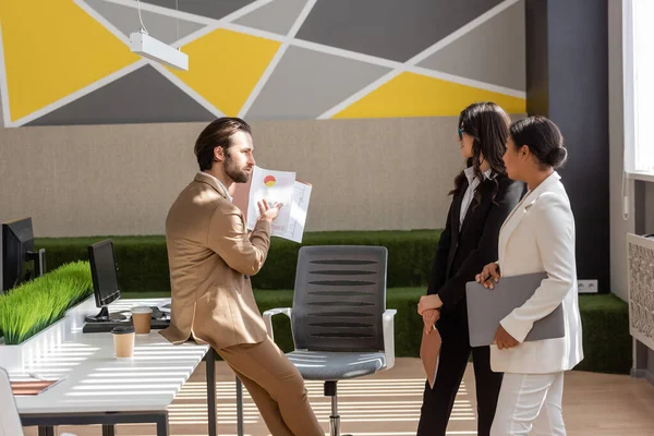 Side view of businessman showing graphs to multicultural colleagues in modern office — Photo de stock