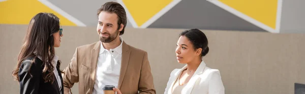Smiling manager with takeaway drink looking at colleague near multiracial businesswoman in office, banner — Stock Photo