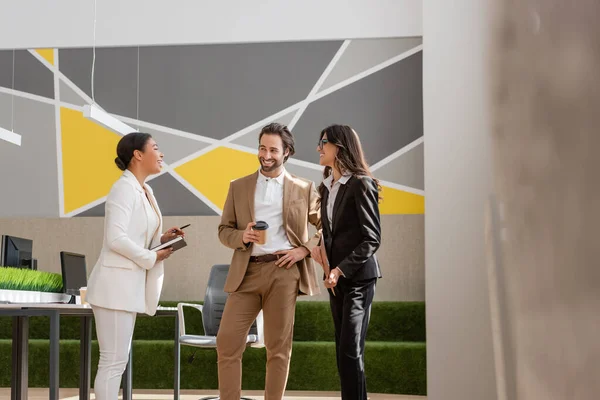 Happy multiethnic managers in formal wear talking near desk with computers in modern office — Stockfoto