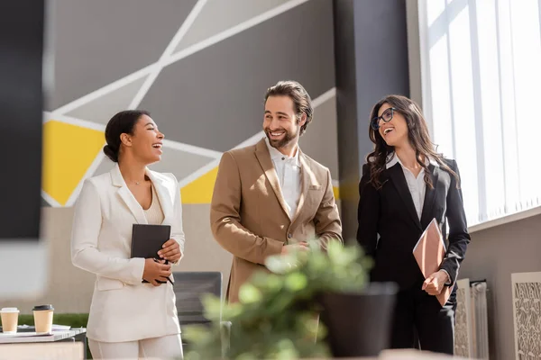 Multiethnic business people in formal wear talking and laughing in office on blurred foreground — Stock Photo