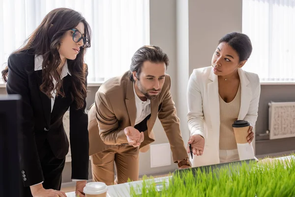 Young manager and multiracial businesswoman with coffee to go pointing at laptop near colleague in eyeglasses in office — Stock Photo