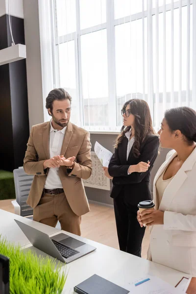 Young manager counting on fingers while talking to multiethnic businesswomen near laptop on desk in office — Stock Photo
