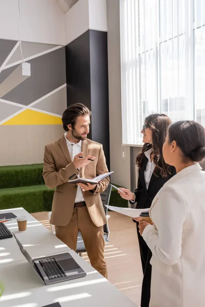 Young manager with notebook talking to multiethnic colleagues near laptop on desk in office — Stock Photo