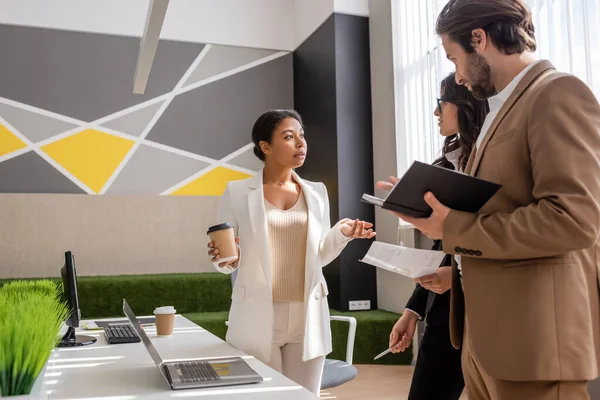 Multiracial businesswoman with paper cup talking to colleagues near desk with computers in office — Stock Photo