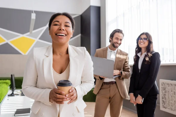 Cheerful multiracial businesswoman with coffee to go looking at camera near colleagues with laptop on blurred background — Stock Photo