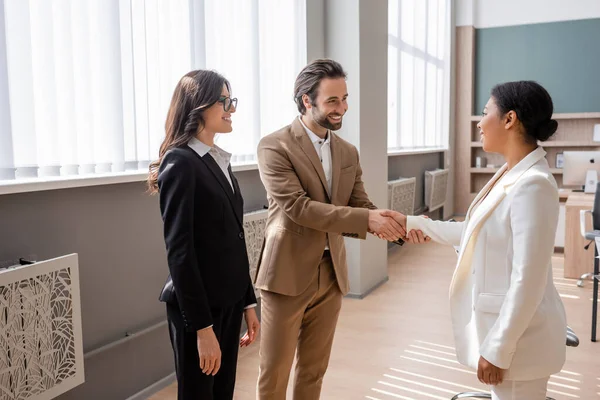 Young and happy manager shaking hands with multiracial businesswoman near colleague in office — Stock Photo