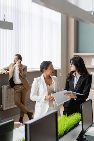 Interracial businesswomen with paper cup and documents talking near blurred manager calling on smartphone in office — Stock Photo