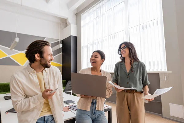 Cheerful multicultural business people with gadgets and documents talking in modern office - foto de stock