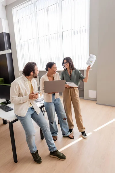 Full length of happy businesswoman showing analytics to interracial colleagues holding laptop and smartphone near desk in office — Foto stock
