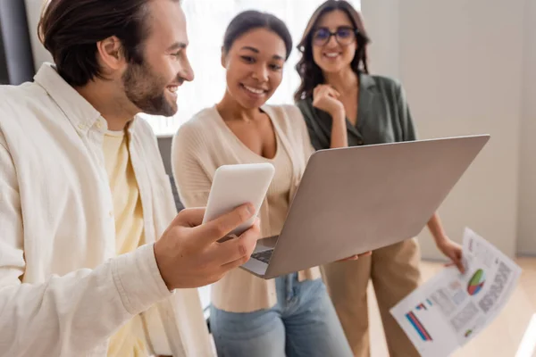 Smiling manager holding smartphone near interracial businesswomen with laptop and documents on blurred background — Photo de stock