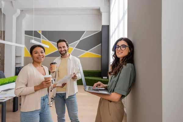 Cheerful multiethnic business colleagues with gadgets and coffee to go looking at camera in office - foto de stock