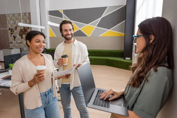 Happy interracial managers with coffee to go looking at colleague standing with laptop in office — Stock Photo