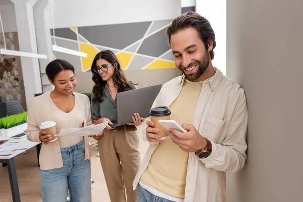 Smiling manager with paper cup using smartphone near interracial businesswomen with documents and laptop — Photo de stock