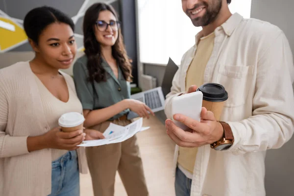 Smiling manager showing smartphone to interracial businesswomen standing with laptop and coffee to go - foto de stock