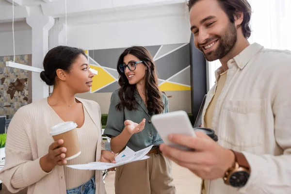 Businesswoman in eyeglasses talking to multiracial colleague near smiling manager with mobile phone - foto de stock