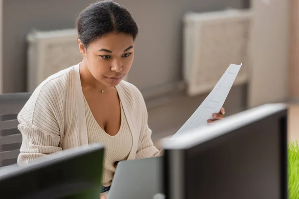 Focused multiracial businesswoman holding document while working on laptop near blurred computer monitors — Stock Photo