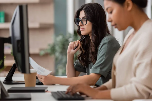 Thoughtful businesswoman in eyeglasses looking at document near multiracial colleague typing on blurred foreground — Stock Photo