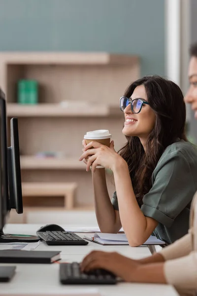Happy manager in eyeglasses sitting with coffee to go near multiracial colleague working on blurred foreground — Stock Photo