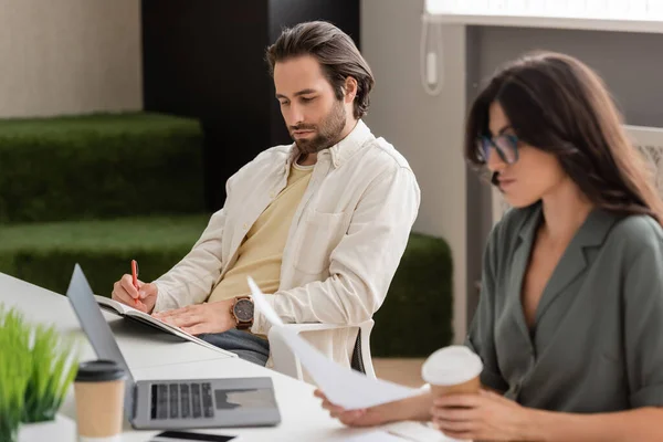 Young manager with notebook and businesswoman with coffee to go sitting near laptop in office — Stock Photo