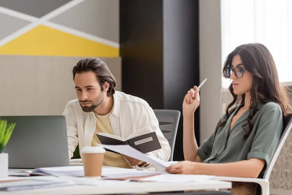 Focused manager with notebook looking at laptop near colleague working with documents in office — Stock Photo