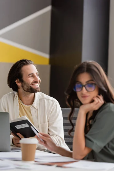 Cheerful businessman with notebook looking away near colleague on blurred foreground — Stock Photo