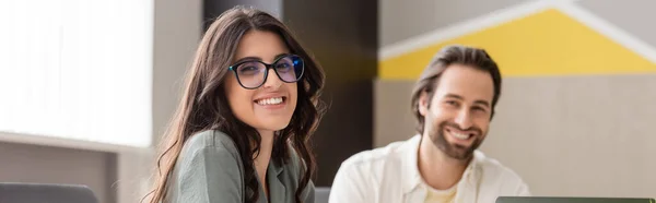 Brunette businesswoman in eyeglasses with blurred bearded colleague smiling at camera in office, banner — Stockfoto