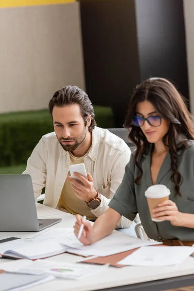 Focused manager with smartphone looking at laptop near colleague holding paper cup while working with documents — Stock Photo