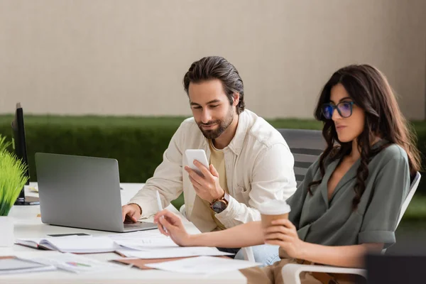 Smiling businessman looking at smartphone and using laptop near blurred colleague with takeaway drink — Stock Photo
