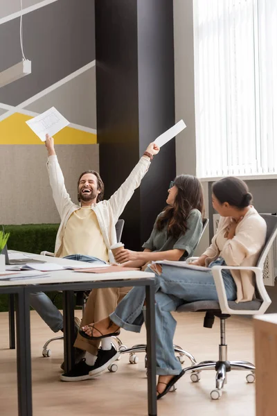 Excited businessman holding documents in raised hands and rejoicing near interracial colleagues sitting in modern office — Fotografia de Stock