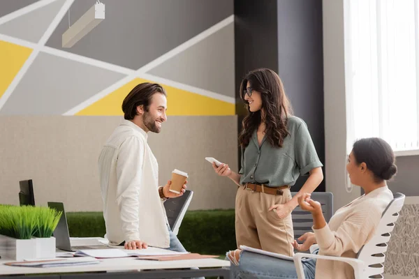 Young manager holding paper cup near smiling colleague with smartphone and multiracial businesswoman sitting in office — Stock Photo