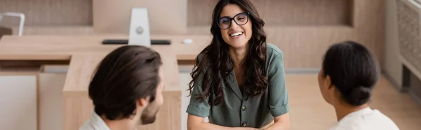 Cheerful businesswoman with long hair and eyeglasses smiling near blurred colleagues in office, banner — Stock Photo