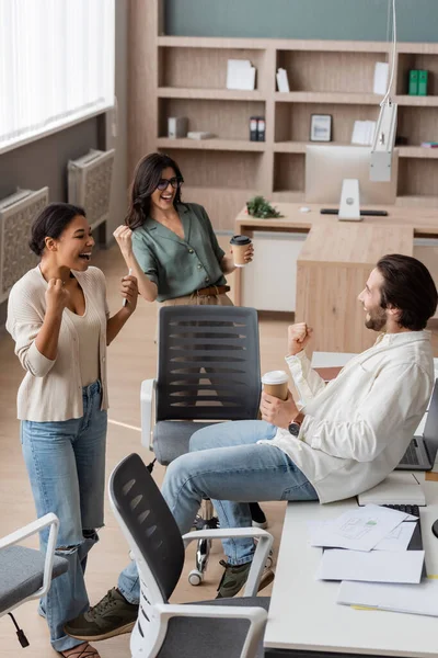 Excited multiethnic business colleagues showing win gesture near desk with documents in office — Foto stock