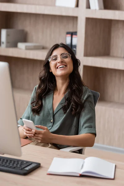 Cheerful businesswoman in eyeglasses holding smartphone and looking at camera near computer monitor and empty notebook — Photo de stock