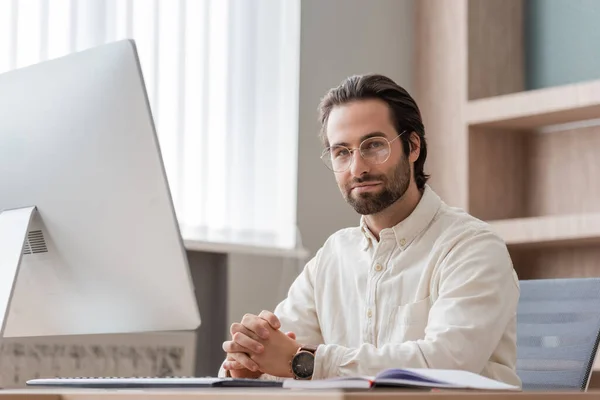 Young bearded businessman in eyeglasses sitting with clenched hands near computer monitor and looking at camera — Photo de stock
