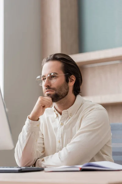 Thoughtful businessman in eyeglasses looking at blurred computer monitor in office — Fotografia de Stock