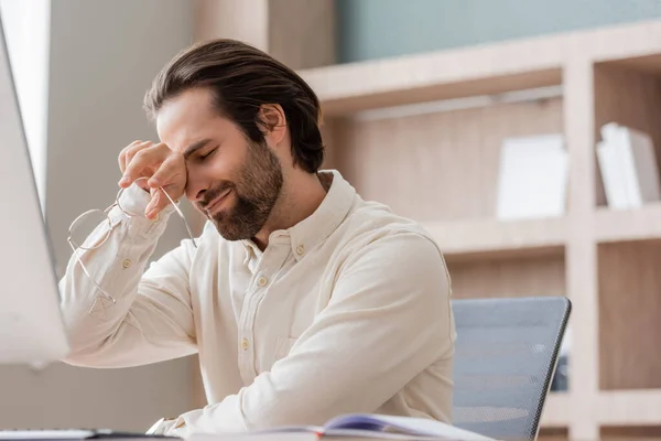 Уставший бизнесмен, держащий очки и вытирающий глаза возле размытого монитора компьютера в офисе — стоковое фото