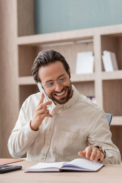 Cheerful bearded businessman in eyeglasses talking on smartphone while looking at notebook on desk — Stock Photo