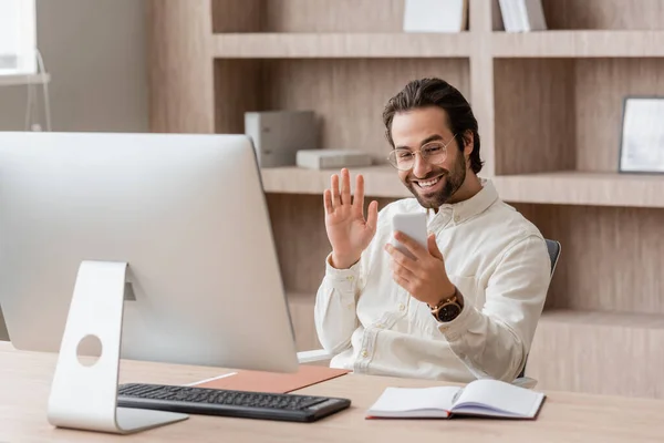 Happy bearded businessman in eyeglasses waving hand during video call on smartphone near computer monitor — Foto stock